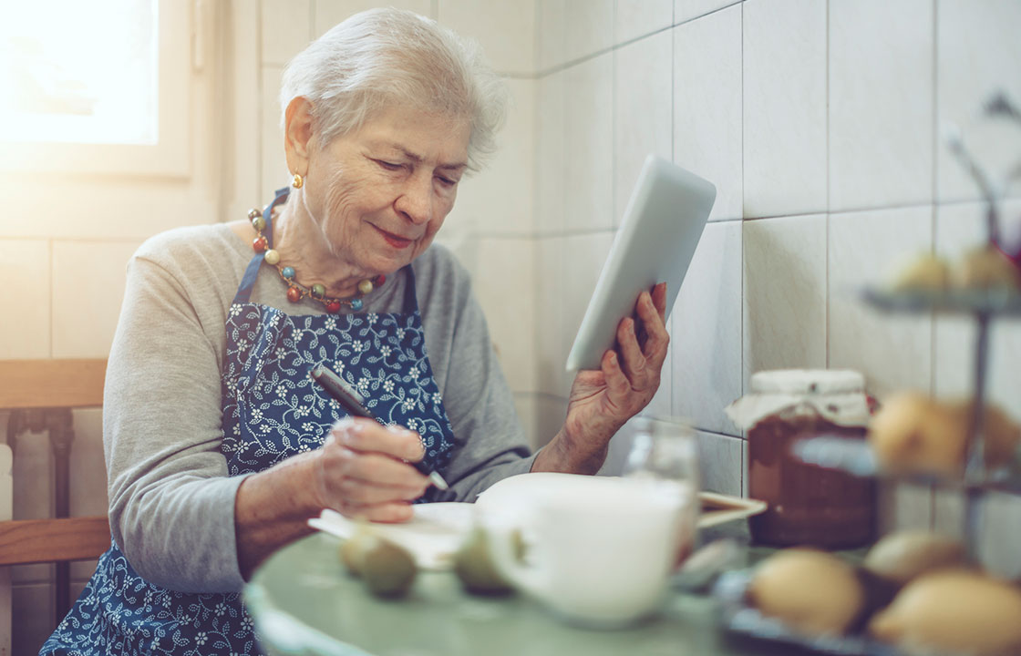 Dame aux cheveux blancs et courts, assise en tablier à une table de cuisine, avec un stylo à la main droite et une tablette à la main gauche.